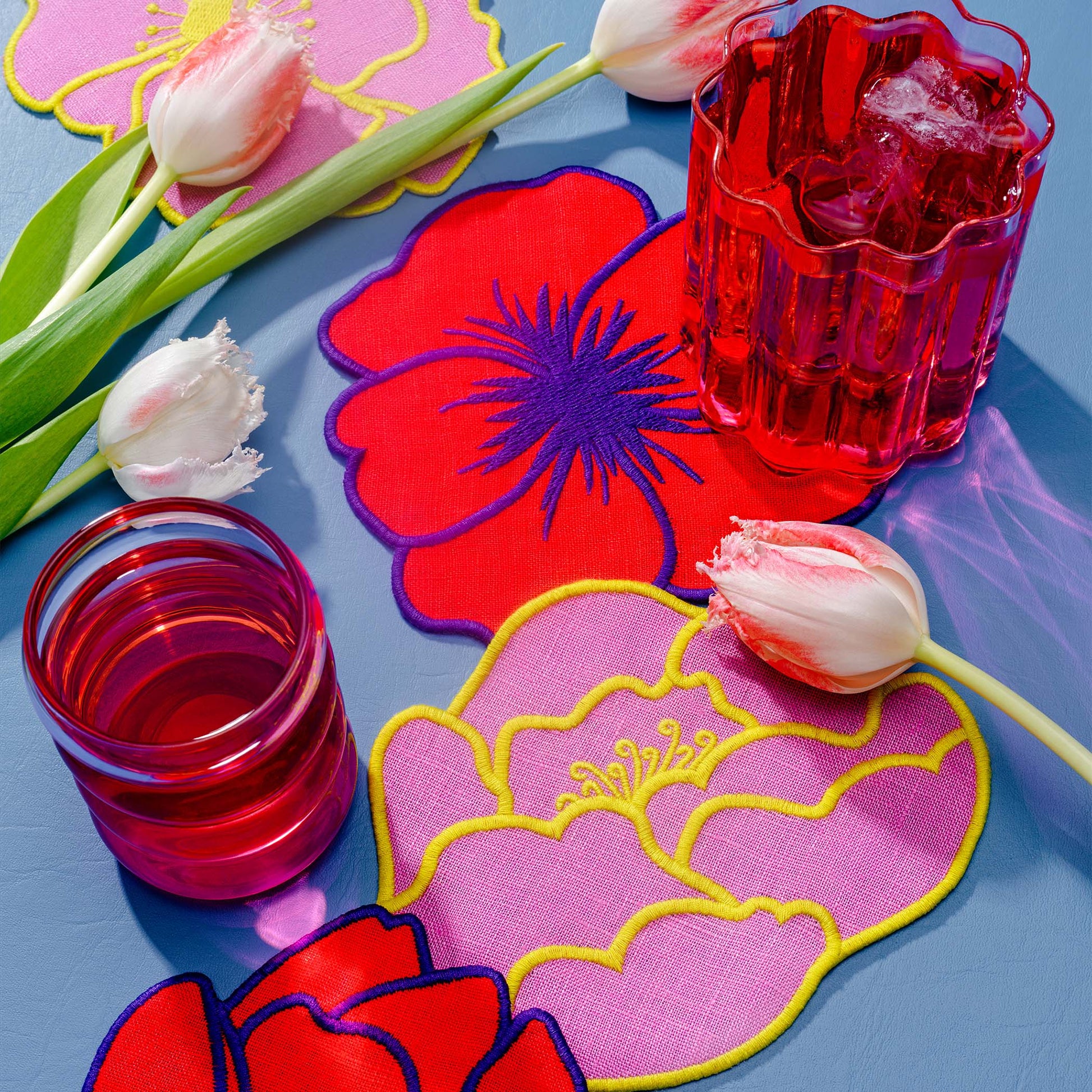 Brightly colored red and pink flower-shaped linen cocktail napkins by Piecework styled with tulips and red drinks on a blue tabletop.