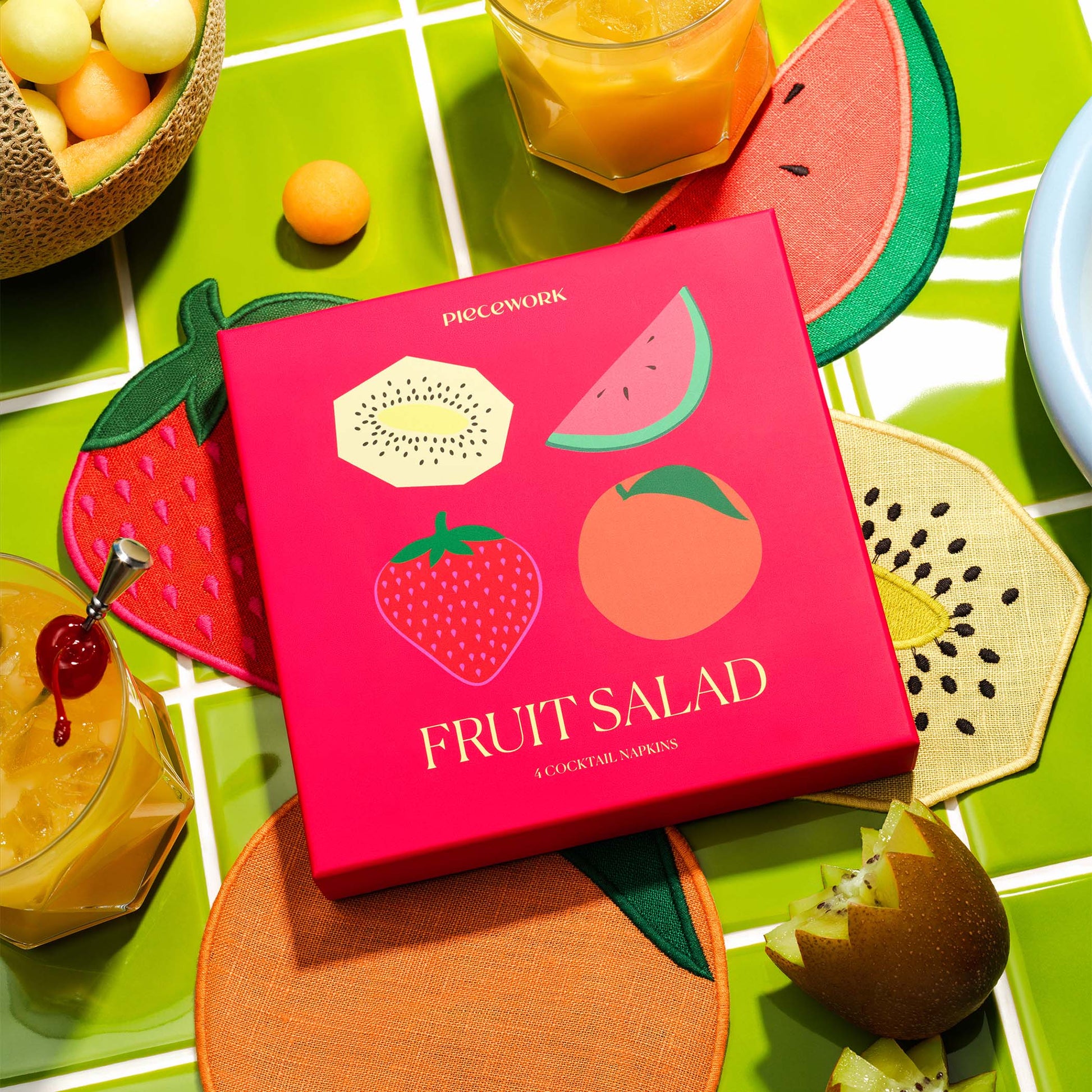 Piecework Fruit Salad Napkins boxed gift set with fruit-shaped linen cocktail napkins styled with drinks and citrus on a green tile backdrop.