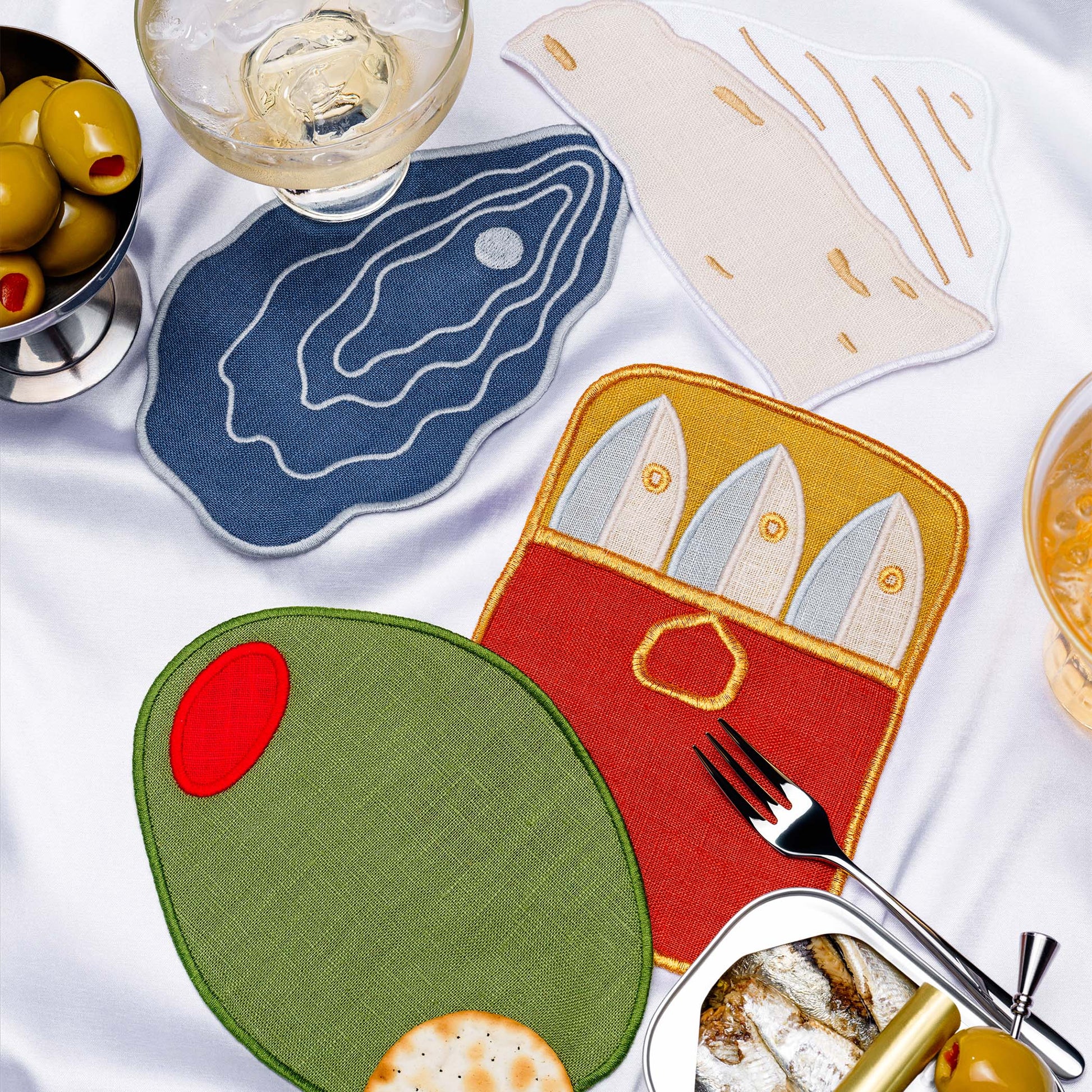 Food-themed cocktail napkins by Piecework, including an olive, a sardine tin, an oyster, and a wedge of cheese, shown styled on a table with drinks and snacks.