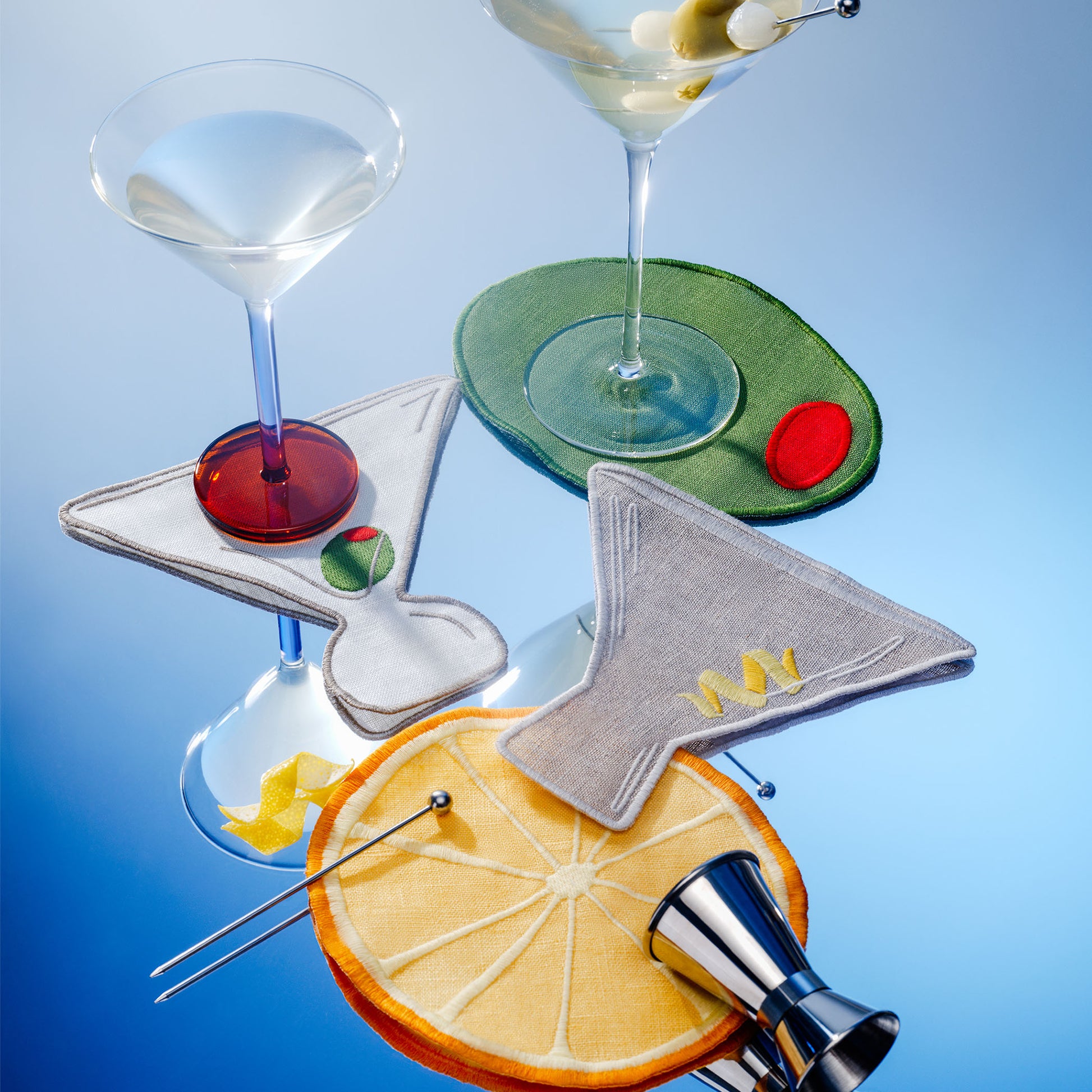 Martini-themed cocktail napkins by Piecework: a dirty martini, a martini with a twist, an olive, and a lemon wheel. Shown styled with drinks, lemon twists, and bar tools.