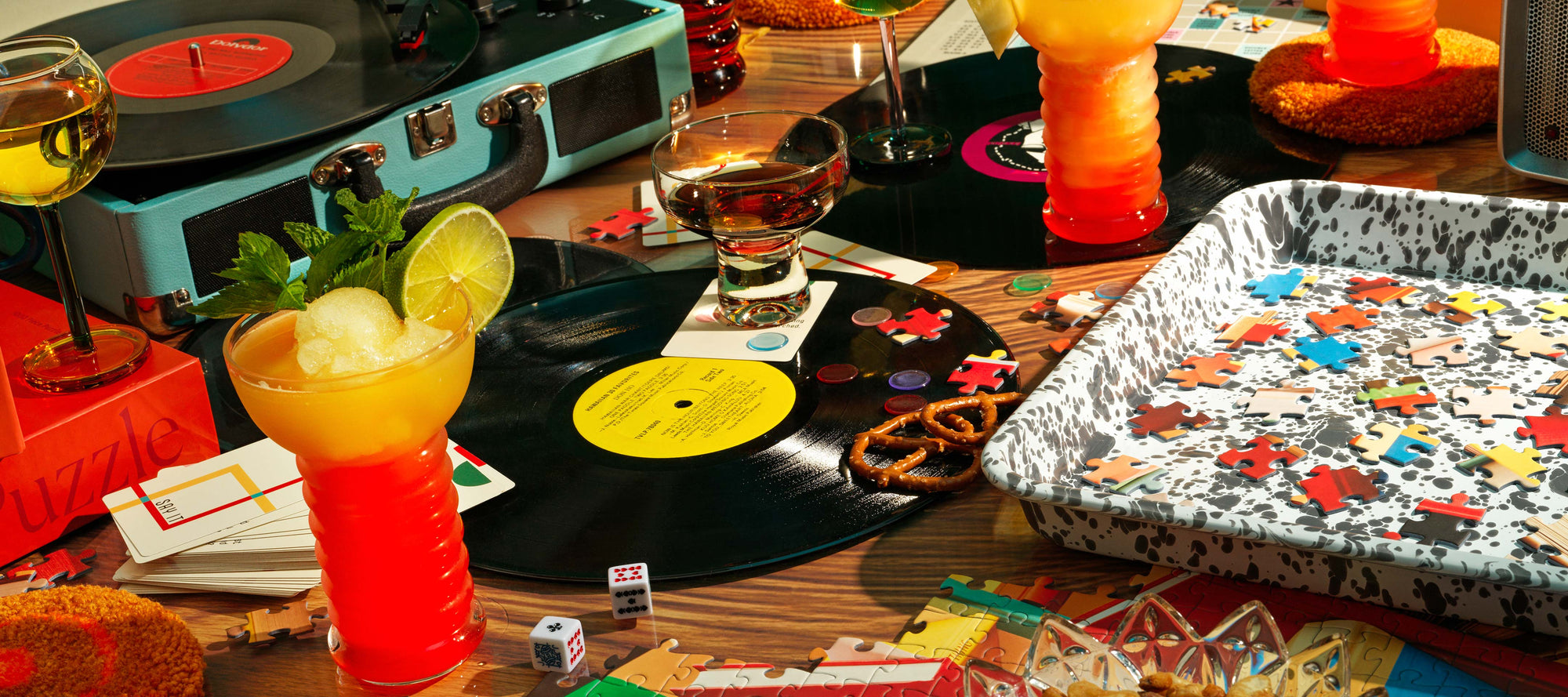 A table scene featuring a record player, cocktails and a puzzle tray filled with puzzle pieces.