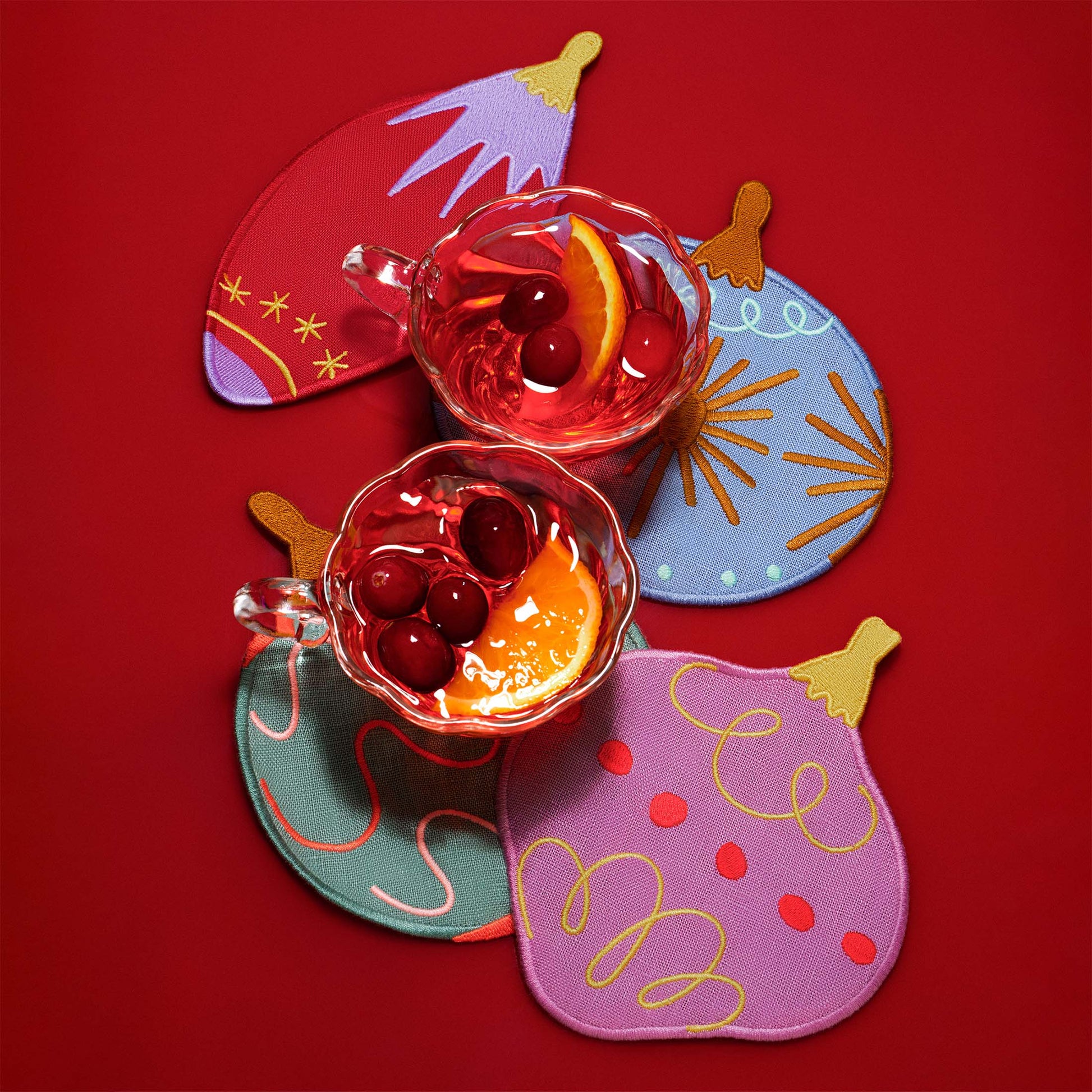 A close-up of ornament-shaped cocktail napkins arranged on a red surface with two glasses of holiday cocktails. The embroidered napkins feature retro-inspired ornament designs in vibrant colors.