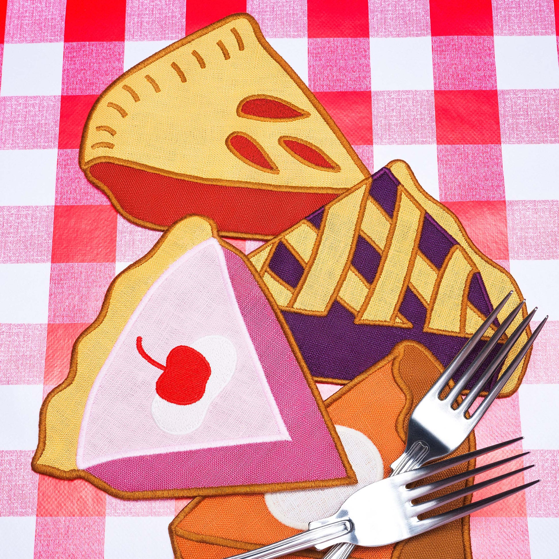 Decorative pie-shaped linen napkins, including one with a cherry on top, on a red and white checkered tablecloth.