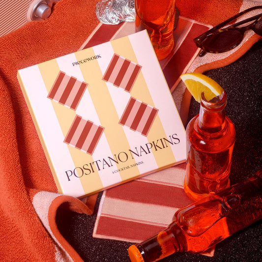 A boxed set of Positano Napkins by Piecework styled on a dark surface with orange towels, a glass bottle of red drink, sunglasses, and an orange garnish. The napkin box has a cream and yellow striped background with diagonal salmon and deep terracotta striped squares.