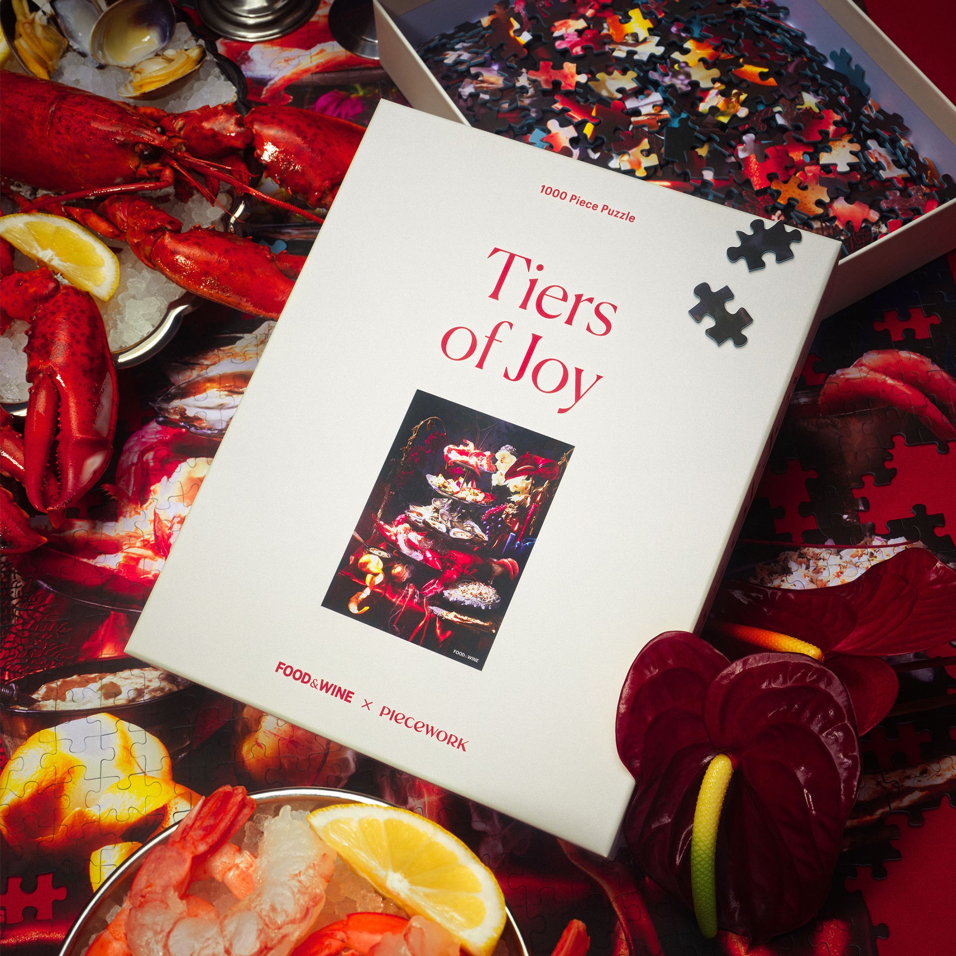 Piecework and Food and Wine's jigsaw puzzle box titled 'Tiers of Joy' surrounded by lobster, florals, shrimp cocktail and puzzle pieces.