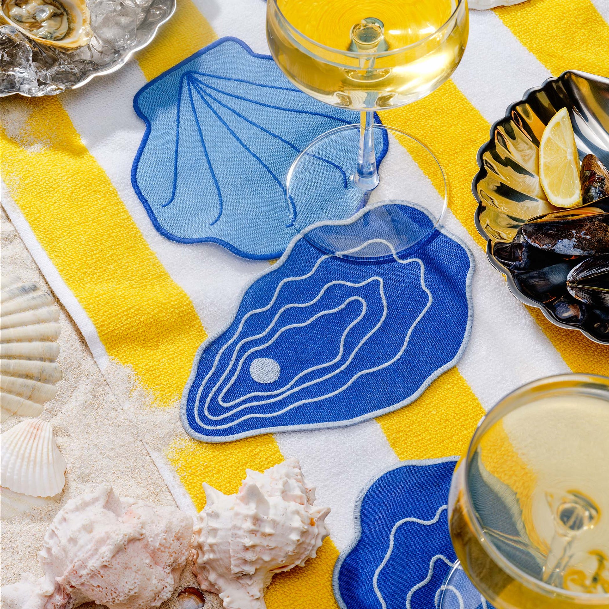 Blue shell-shaped cocktail napkins by Piecework styled with wine, mussels, and seashells on striped beach towel.