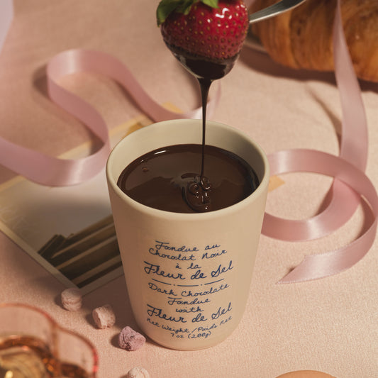 An image of a strawberry being dipped in the dark chocolate fondue on a pink table top.