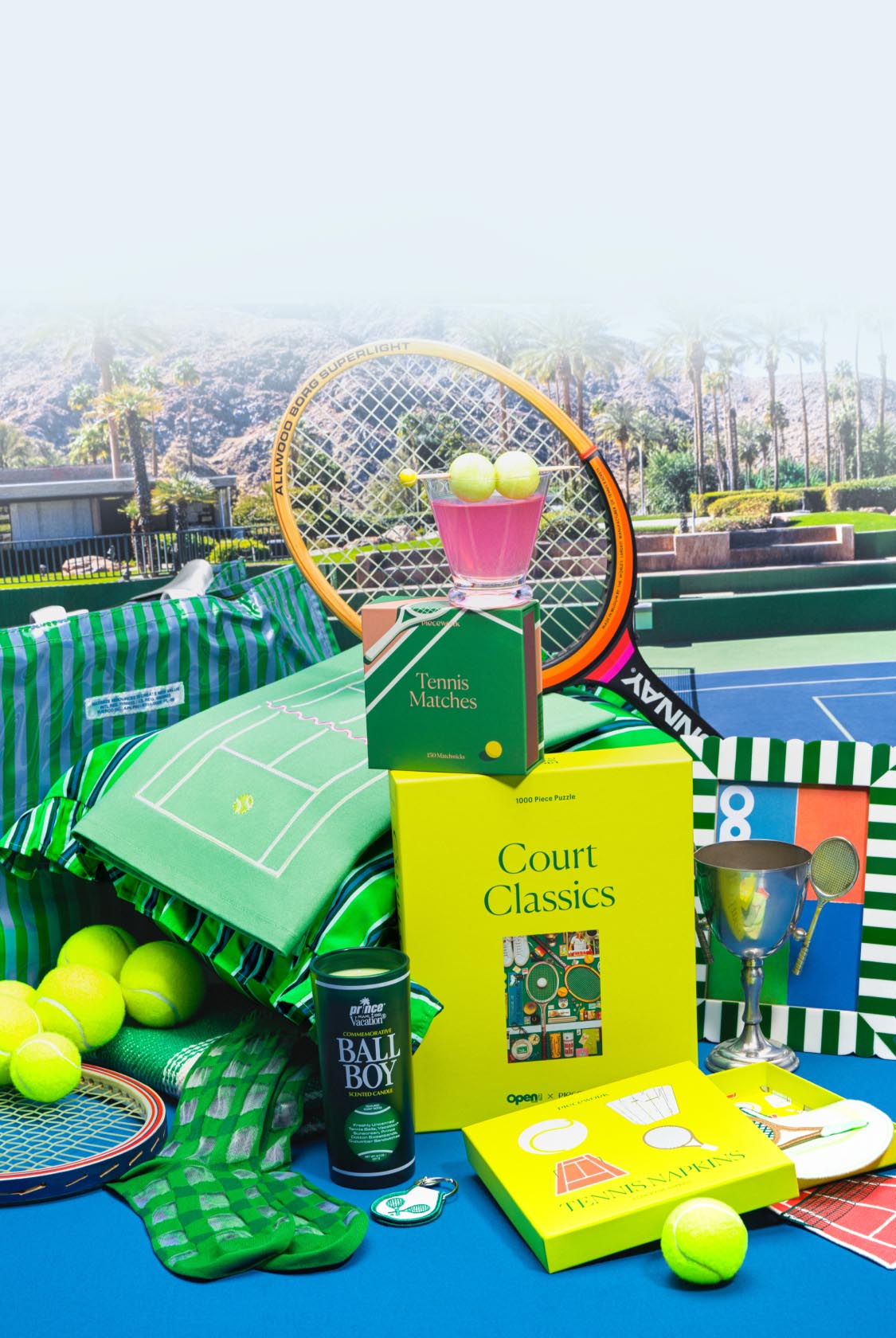 Tennis-themed product grouping including rackets, tennis balls, tennis cocktail napkins, tennis matches, and a tennis puzzle shown outdoors on a court.