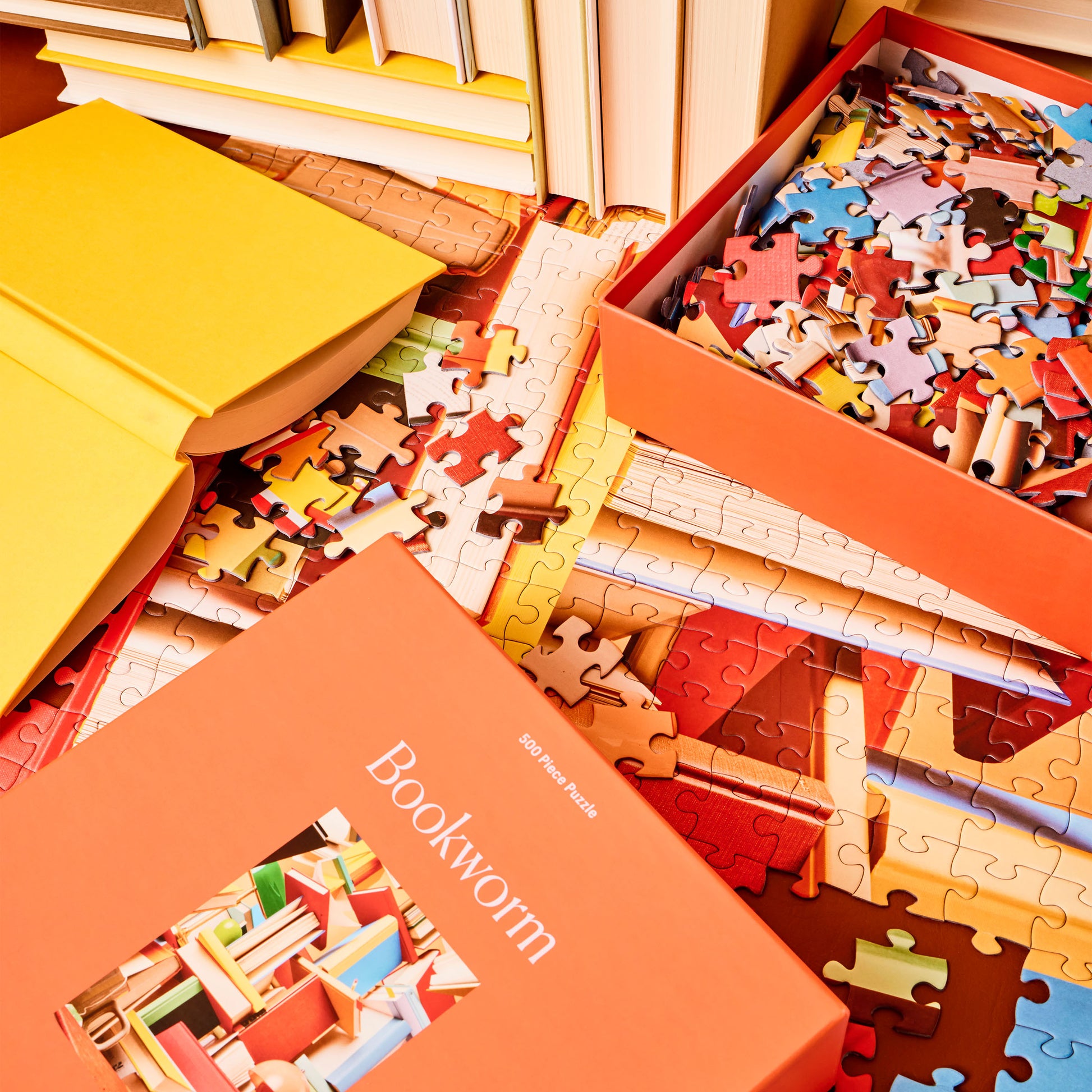Box for Bookworm, a 500-Piece Puzzle from Piecework Puzzles, placed in a scene of books, a box of puzzle pieces, and the partially completed Bookworm puzzle.
