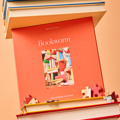 Box for Bookworm, a 500-Piece Puzzle from Piecework Puzzles, placed in a stack of books with scattered puzzle pieces.
