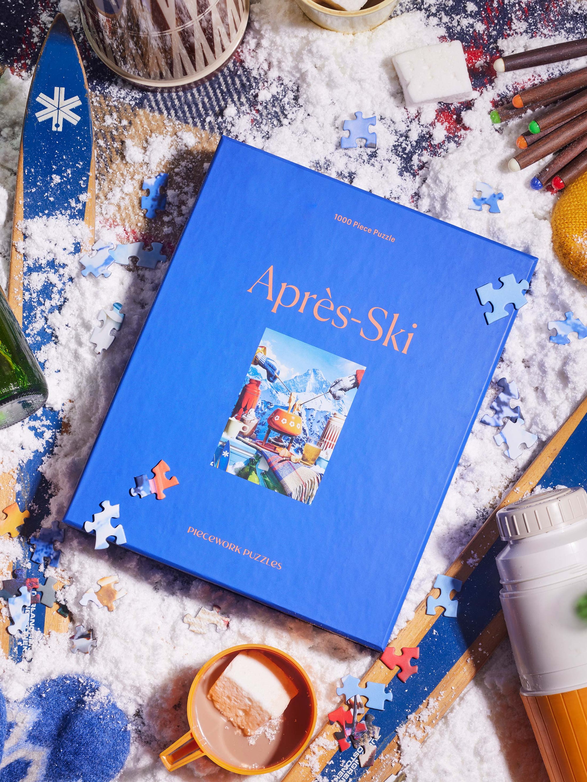 Après-Ski 1000-piece puzzle in a cozy winter scene, featuring a bold blue box on a snow-dusted backdrop with hot cocoa, marshmallows, and ski-themed details.