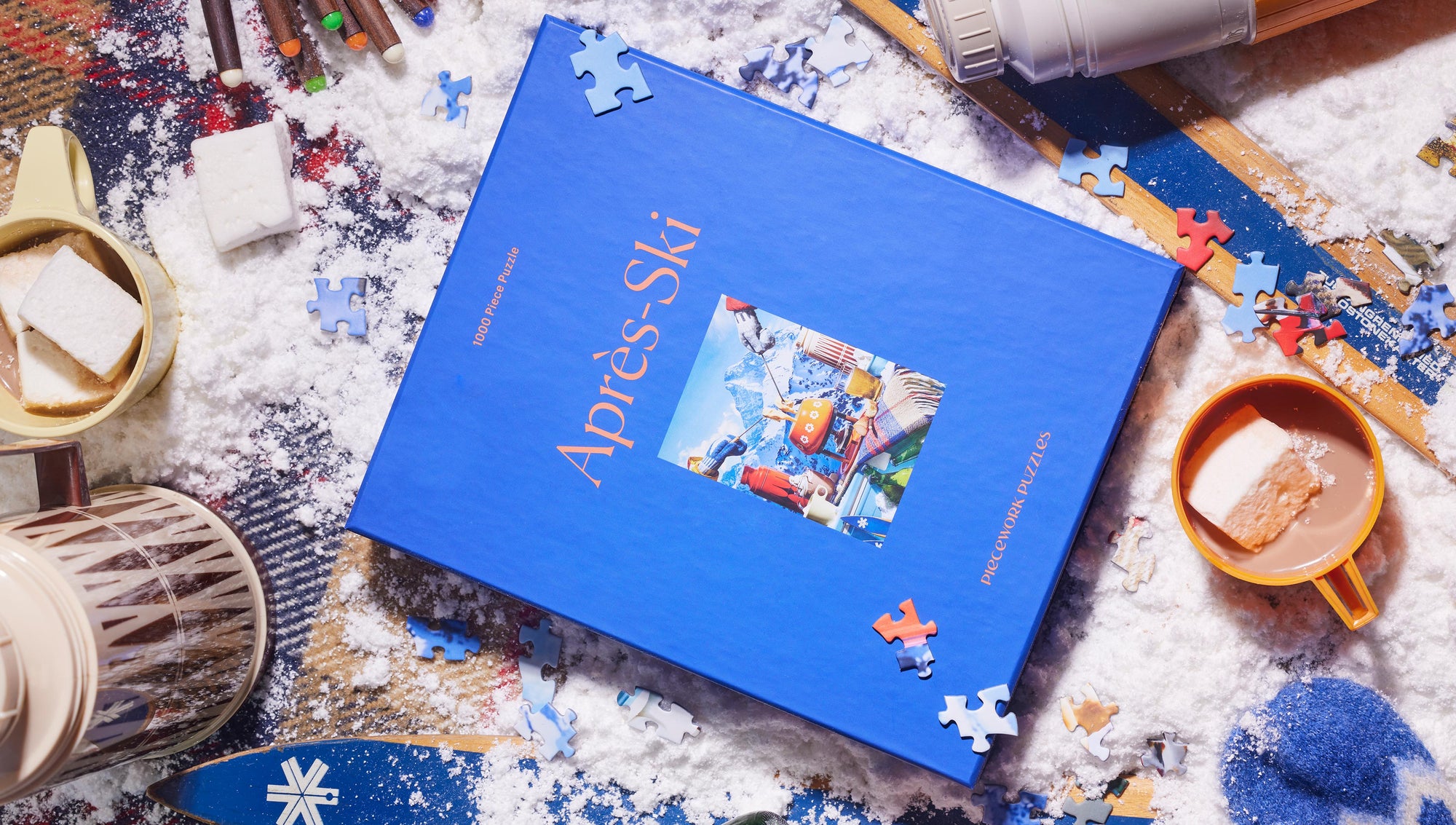 Après-Ski 1000-piece puzzle in a cozy winter scene, featuring a bold blue box on a snow-dusted backdrop with hot cocoa, marshmallows, and ski-themed details.