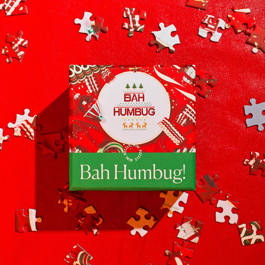 A festive red and green scene featuring the “Bah Humbug!” mini puzzle by Piecework Puzzles. The puzzle box, decorated with a cross-stitched holiday motif reading “BAH HUMBUG,” sits on a bright red surface surrounded by scattered puzzle pieces. Playful and cheeky, it captures a humorous twist on classic Christmas spirit.