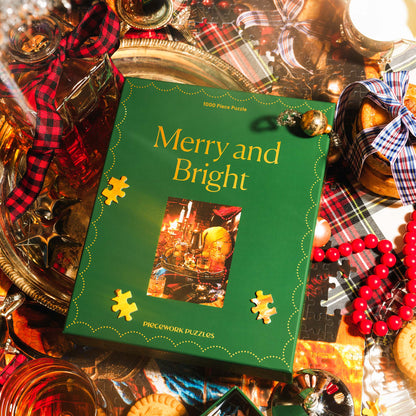 Piecework Puzzles' 1000-piece puzzle Merry and Bright photographed in a scene is filled with flickering candlesticks, tinsel and tartan. The box is photographed top-down, laying flat on a table.