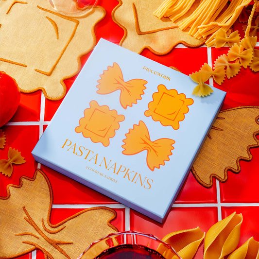 Piecework Pasta Napkins boxed gift set with ravioli and farfalle-shaped linen cocktail napkins styled with pasta on a red tile backdrop.