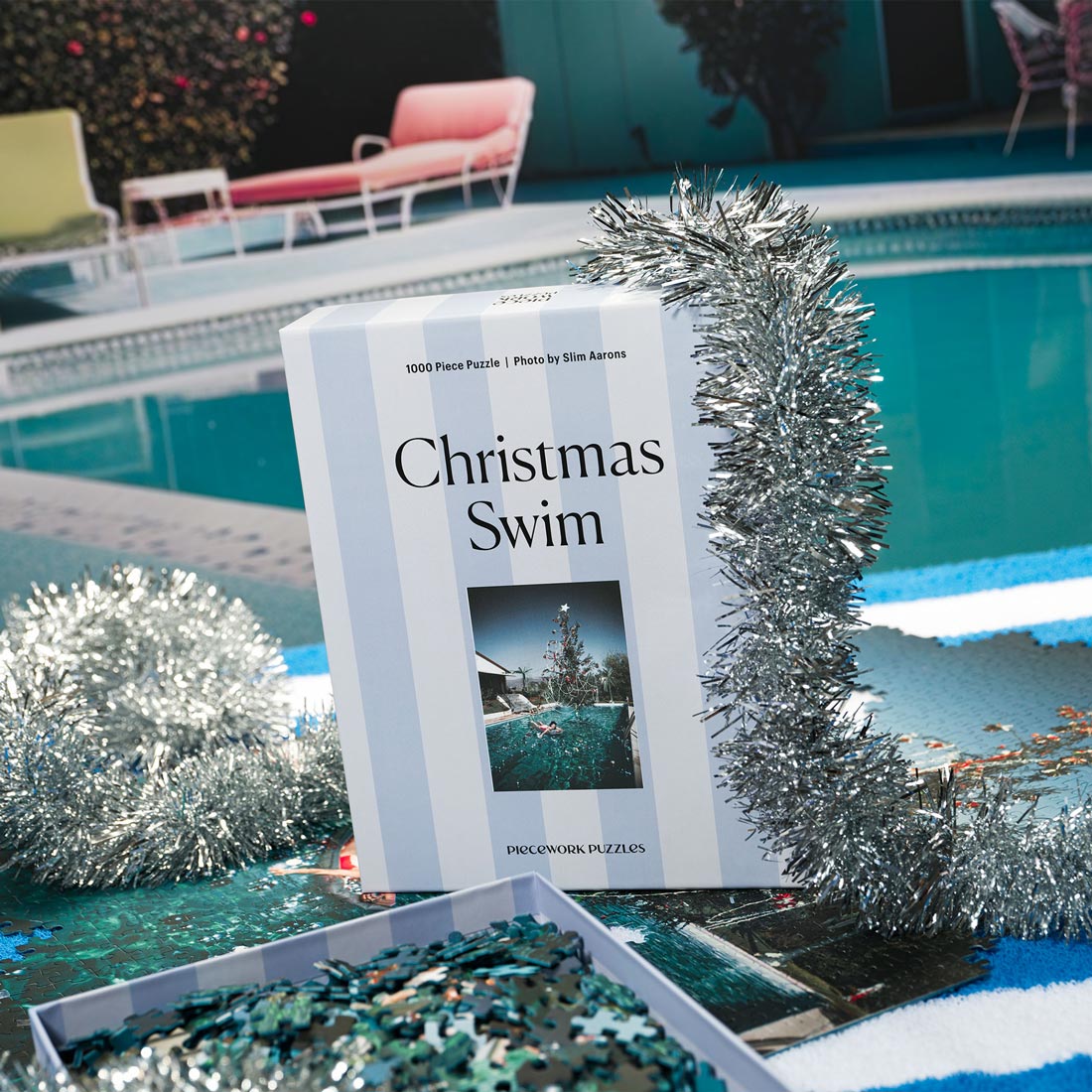 A puzzle box titled 'Christmas Swim' by Slim Aarons, depicting a photograph of a woman floating in a pool with a festive, holiday-themed decoration in the foreground.