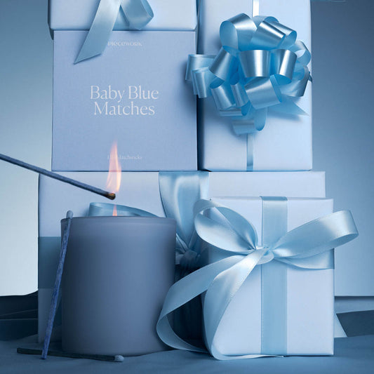 Blue candle and matches surrounded by blue gift boxes and ribbons on a matching background. The focal point of the image shows a match lighting a candle and the packaging for Piecework's Baby Blue Matches.