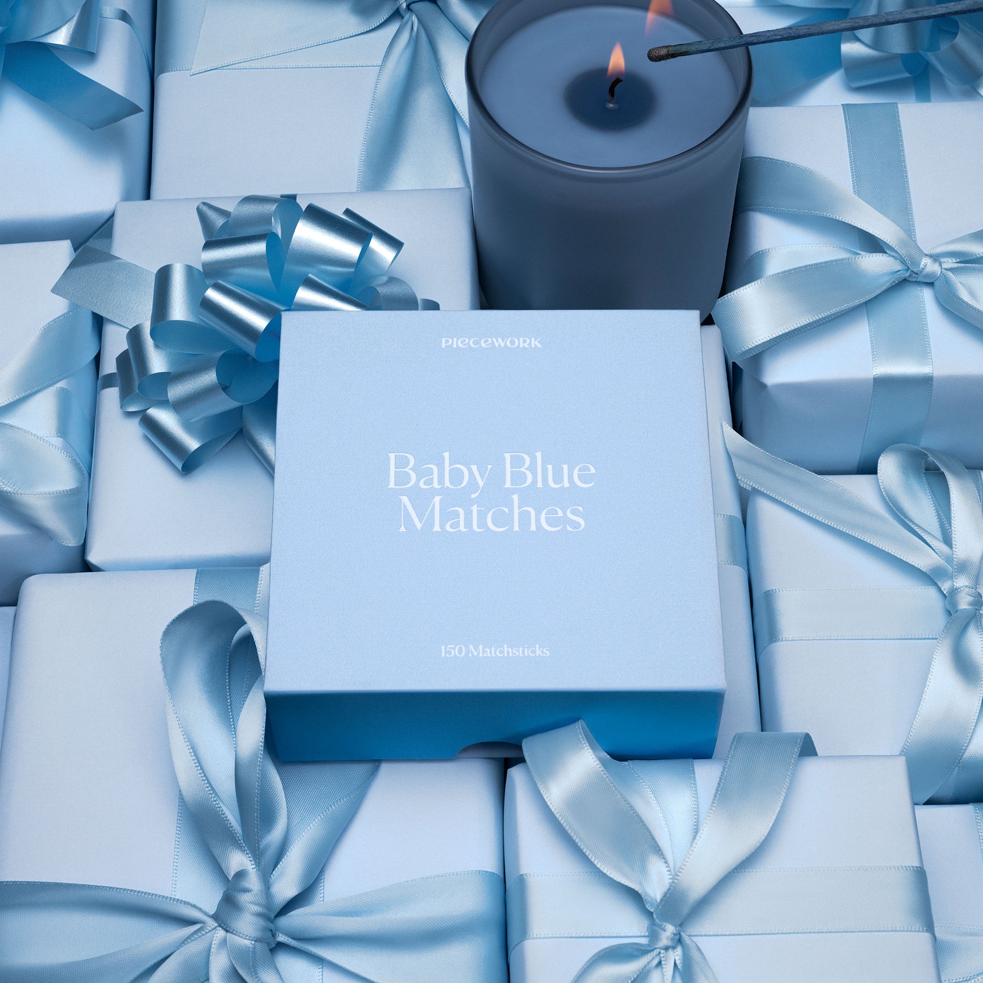 Blue candle and matches surrounded by blue gift boxes and ribbons on a matching background. The focal point of the image is the packaging for Piecework's Baby Blue Matches.