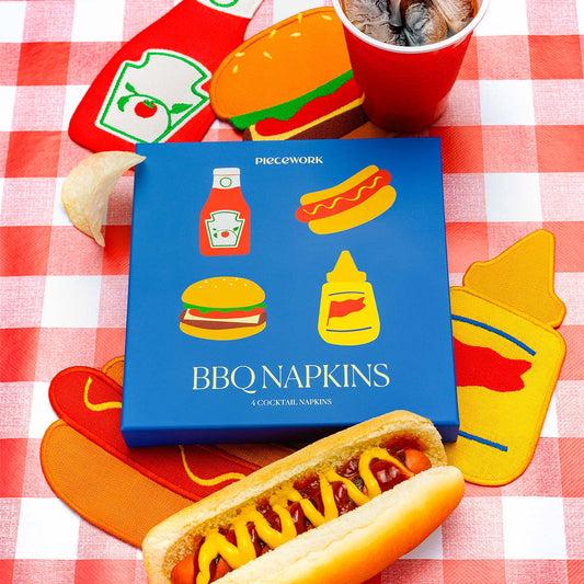 BBQ-themed linen cocktail napkin set by Piecework featuring ketchup, mustard, burger, and hot dog designs on a red gingham picnic table with chips and drinks.