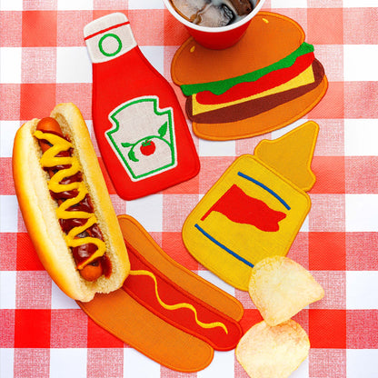 Colorful embroidered cocktail napkins shaped like ketchup, mustard, a hamburger, and a hot dog styled on a red gingham tablecloth beside snacks and soda.