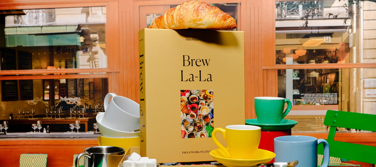 Brew La-La 1000-piece puzzle box topped with a croissant, surrounded by colorful espresso cups at a café window