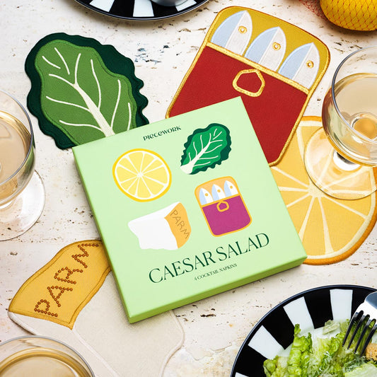 A bright tabletop scene featuring the “Caesar Salad” cocktail napkins set by Piecework. The green box displays playful embroidered motifs—a lemon slice, lettuce leaf, wedge of parmesan, and anchovy tin—matching the four linen napkins arranged around it.