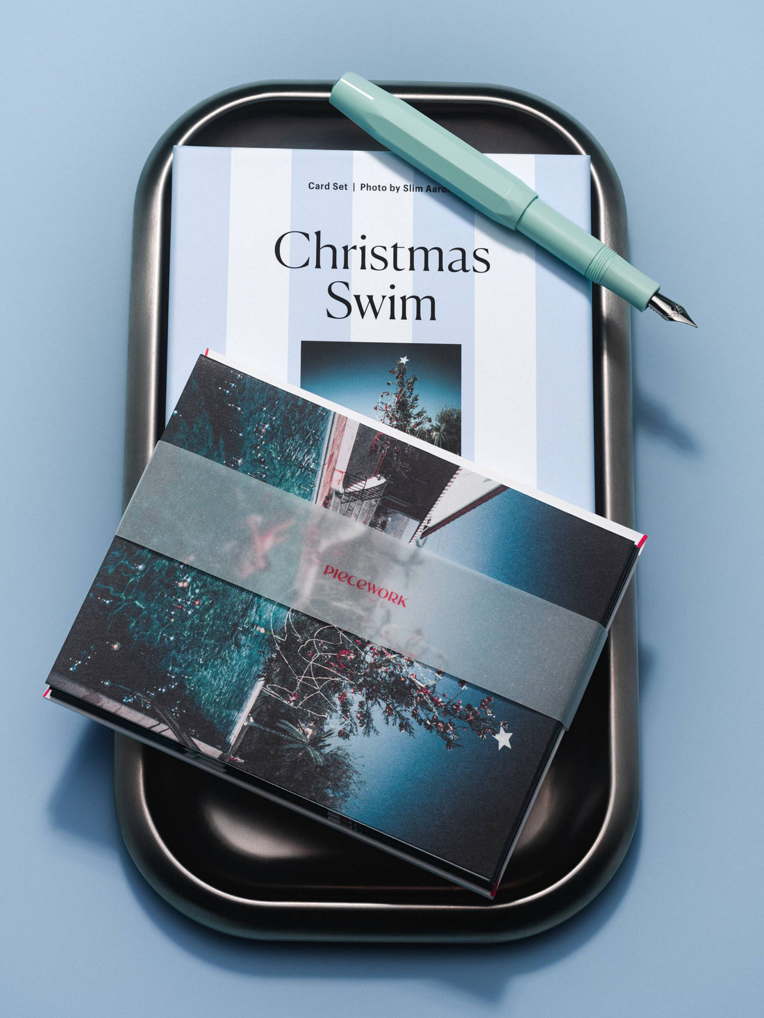 Piecework Christmas Swim boxed notecard set with a vellum-banded card and mint fountain pen on a dark tray, blue background