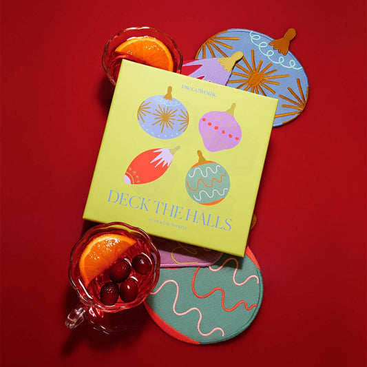A vibrant green box labeled “Deck the Halls” by Piecework is surrounded by four ornament-shaped embroidered linen cocktail napkins in rich jewel tones. Each napkin features a unique retro-inspired design. Two glasses of festive drinks are adorned with orange slices and cherries are placed around the napkins on a bright red background.