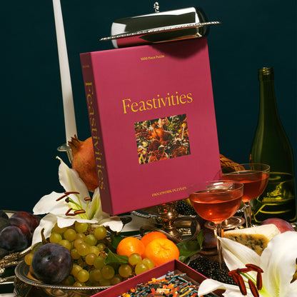 The Feastivities 1000-piece puzzle box stands upright on a holiday table surrounded by fresh fruit, white lilies, candles, wine glasses, and silver servingware. A box of puzzle pieces with rich autumnal colors is visible below.