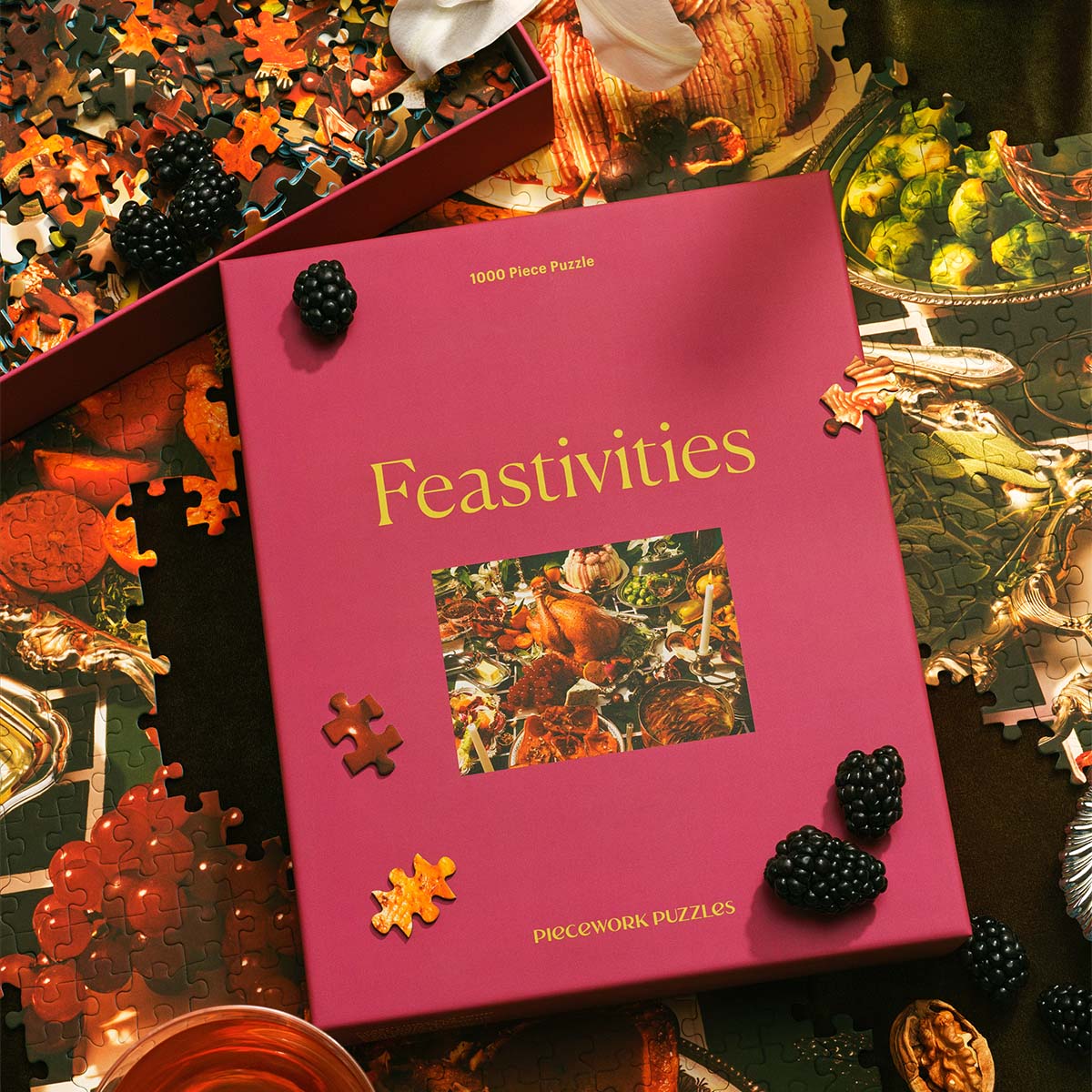 The Feastivities puzzle box lies flat on a table, surrounded by scattered puzzle pieces and blackberries. A partially completed section of the puzzle shows a festive spread of holiday food.