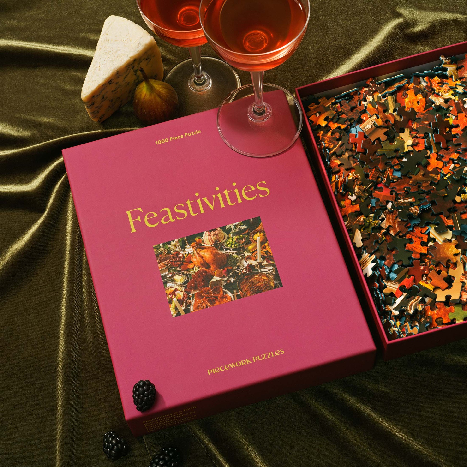 The Feastivities puzzle box sits next to a wine glass, wedge of cheese, and a box of puzzle pieces on dark green velvet. A cozy and indulgent holiday setup.