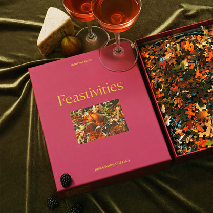 The Feastivities puzzle box sits next to a wine glass, wedge of cheese, and a box of puzzle pieces on dark green velvet. A cozy and indulgent holiday setup.