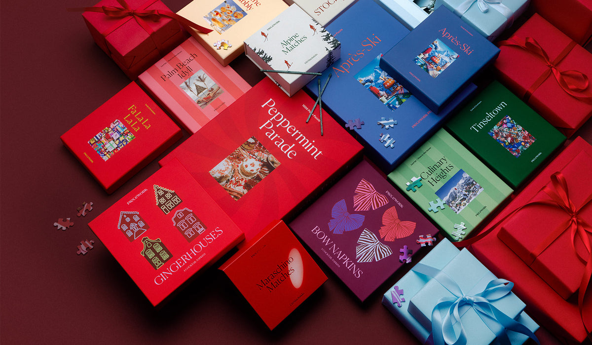 The Piecework Holiday Collection laid out among red and pale blue wrapped gifts on a deep burgundy background. It includes the Peppermint Parade and Après-Ski 1000-piece puzzles, the Tinseltown, Fa La La La La, Palm Beach Idyll, Culinary Heights and Après-Ski stationery sets, Gingerhouses and Bow cocktail napkins, and Maraschino and Alpine matches, with puzzle pieces scattered around.