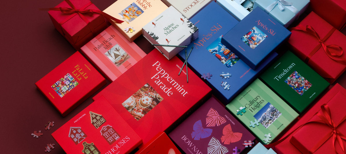 The Piecework Holiday Collection laid out among red and pale blue wrapped gifts on a deep burgundy background. It includes the Peppermint Parade and Après-Ski 1000-piece puzzles, the Tinseltown, Fa La La La La, Palm Beach Idyll, Culinary Heights and Après-Ski stationery sets, Gingerhouses and Bow cocktail napkins, and Maraschino and Alpine matches, with puzzle pieces scattered around.