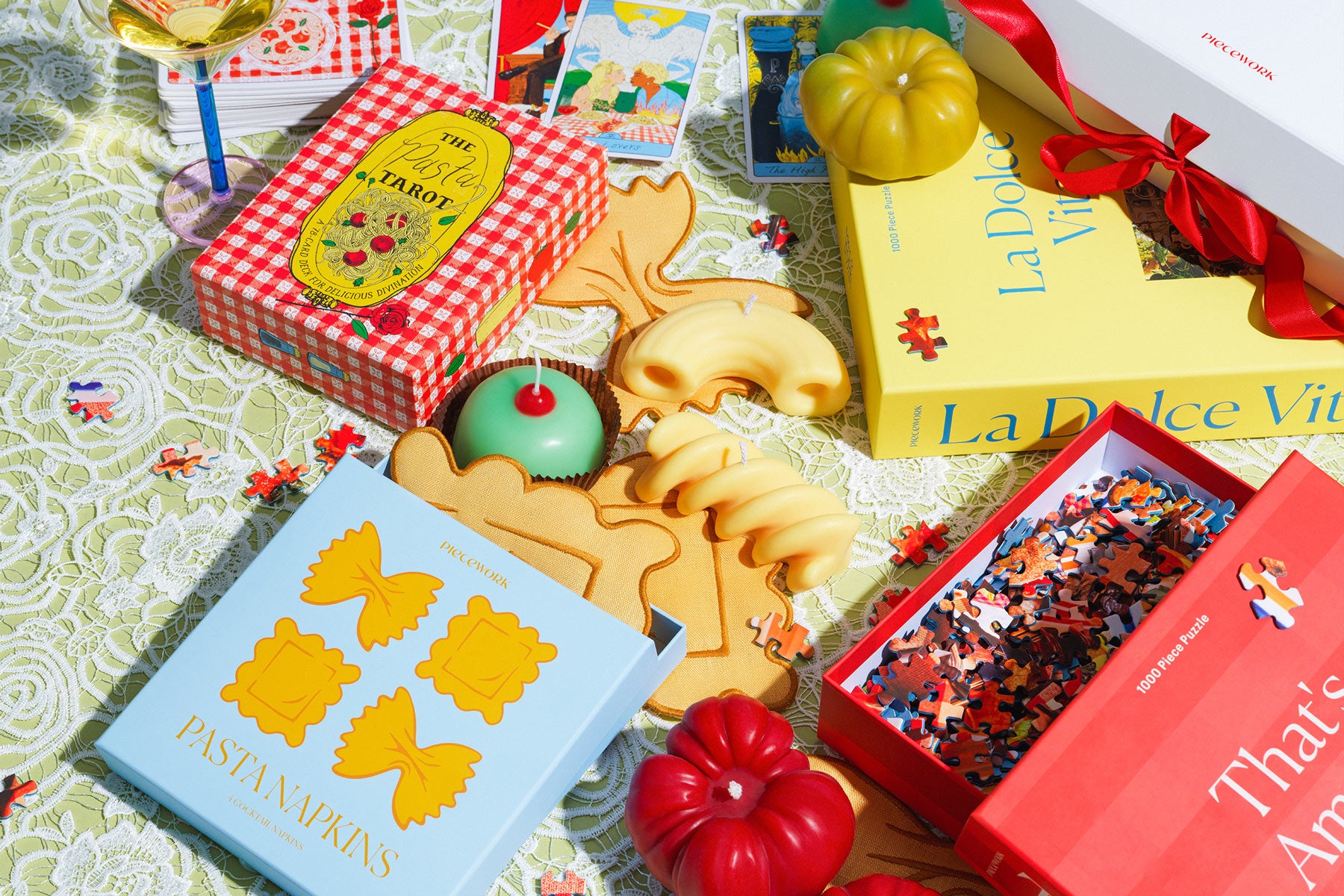 A colorful Piecework scene: “Pasta Tarot,” “That’s Amore,” “La Dolce Vita,” and “Pasta Napkins” on lace with tarot cards, tomato and pasta candles, puzzle pieces, a martini glass, and a red ribbon — a whimsical Italian-inspired picnic moment.