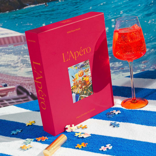 Pink 'L'Apéro' puzzle box with an aperol spritz cocktail and scattered puzzle pieces on a blue and white towel by a beach.