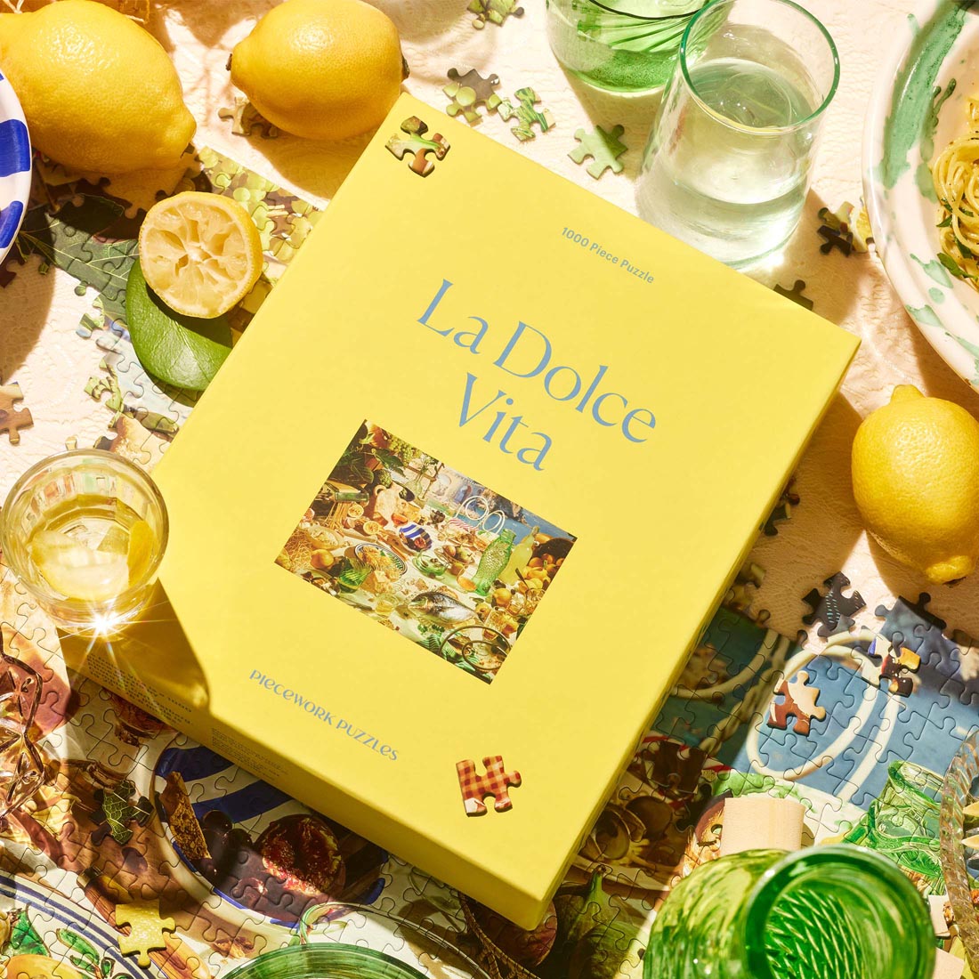A sunlit table scene featuring the “La Dolce Vita” 1000-piece jigsaw puzzle by Piecework Puzzles. The bright yellow box rests among scattered puzzle pieces, fresh lemons, glasses of water and wine, and a plate of pasta.
