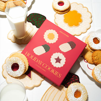 A red box labeled “Holiday Cookies” by Piecework is surrounded by jam-filled Linzer-style cookies and four embroidered linen cocktail napkins shaped like cookies. The napkins feature scalloped edges, powdered sugar stitching, and jammy centers. A glass of milk sits beside the cookies and napkins on a white tabletop.