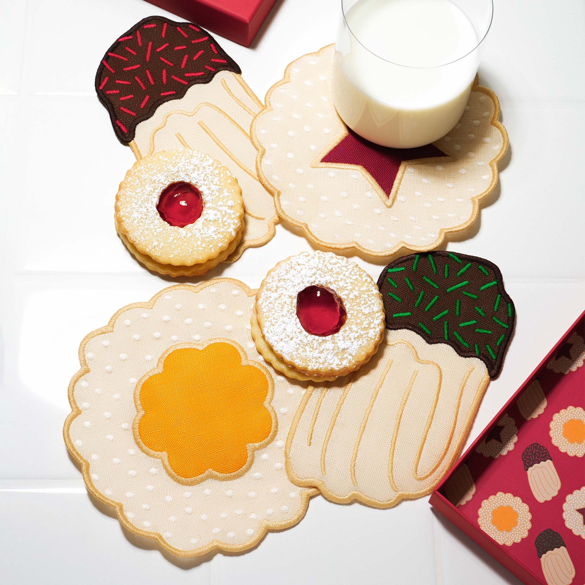 Four embroidered cookie-shaped linen cocktail napkins arranged on a white surface, surrounded by Linzer-style cookies and a glass of milk. Each napkin resembles a different cookie with scalloped edges and jam-like centers, evoking a festive Christmas cookie party.