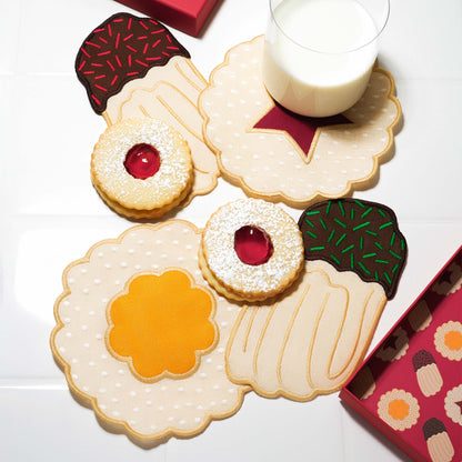 Four embroidered cookie-shaped linen cocktail napkins arranged on a white surface, surrounded by Linzer-style cookies and a glass of milk. Each napkin resembles a different cookie with scalloped edges and jam-like centers, evoking a festive Christmas cookie party.