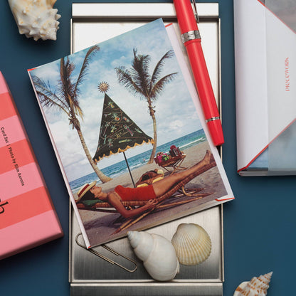 Piecework Palm Beach Idyll Stationery Set: Slim Aarons sunbather notecard on a silver tray with a red pen and shells, navy