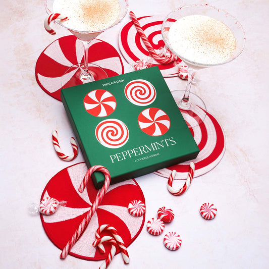 A green box labeled “Peppermints” by Piecework sits surrounded by four red-and-white embroidered linen cocktail napkins, each designed to resemble peppermint candies. The scene is styled with real candy canes, striped peppermint sweets, and two martini glasses filled with creamy cocktails on a light surface. The overall look evokes a festive, candy-striped holiday bar.