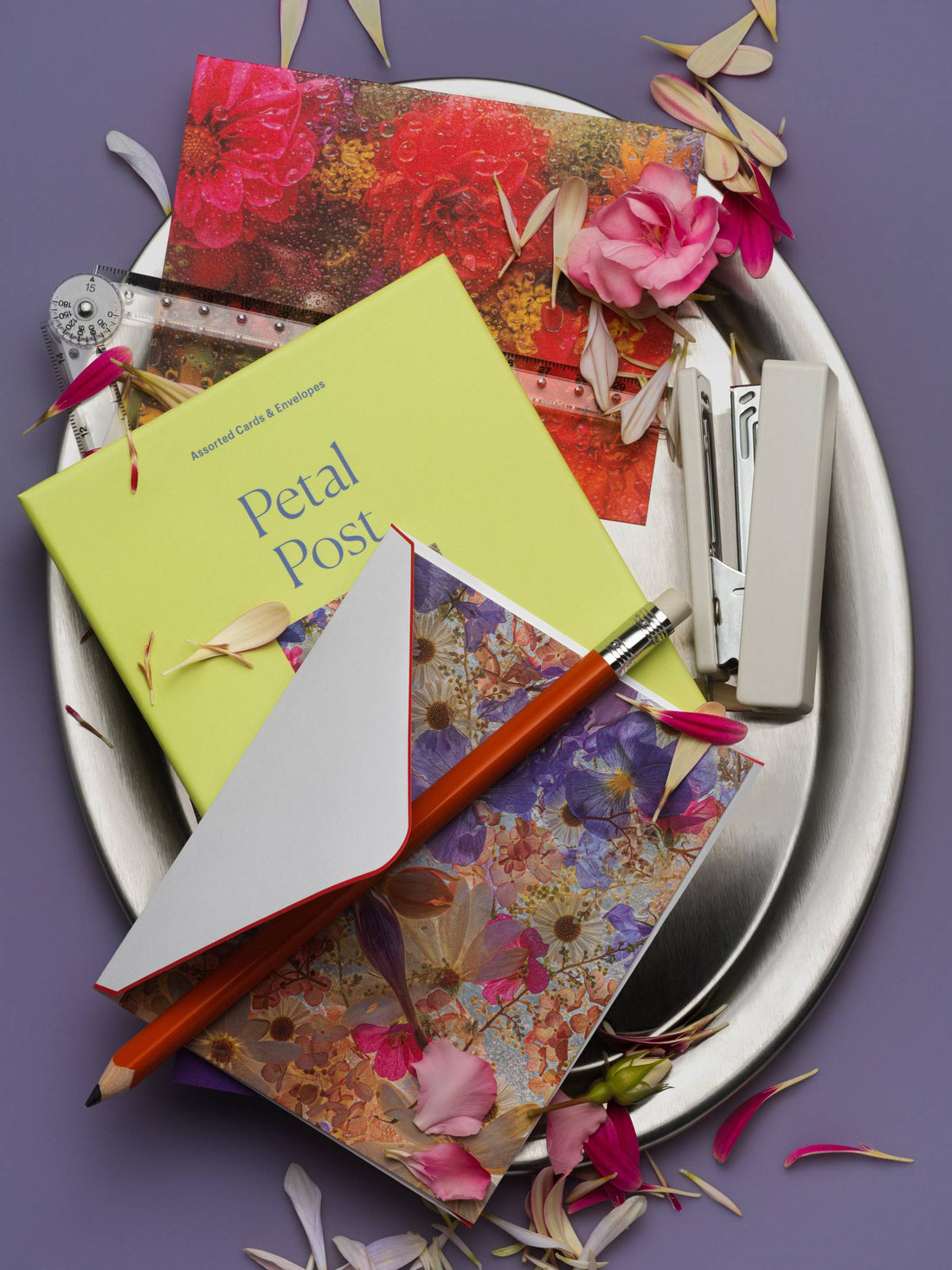 Piecework Petal Post notecards and box on a silver tray with rose petals, orange pencil and stapler on slate purple