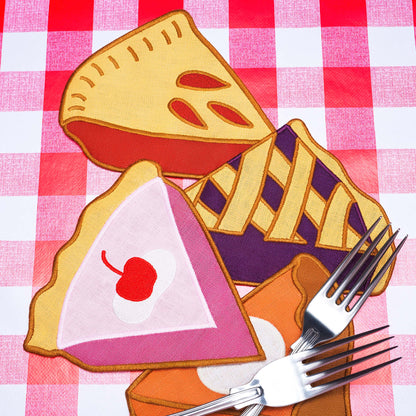 Decorative pie-shaped linen napkins, including one with a cherry on top, on a red and white checkered tablecloth.