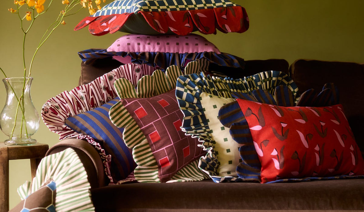 Piecework petal and ruffle pillows piled on a brown velvet sofa beside a vase of yellow orchids
