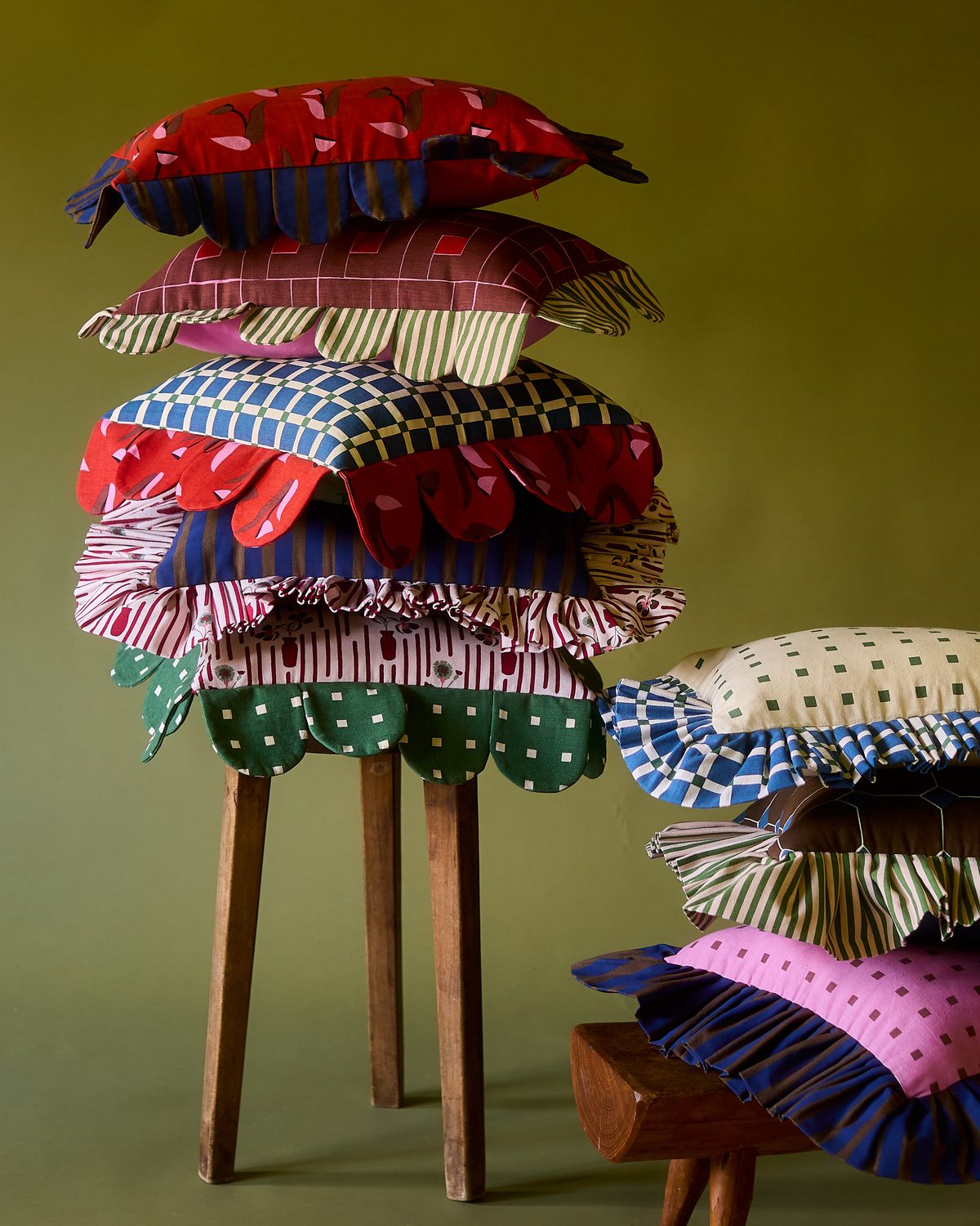 Piecework petal, ruffle and scalloped pillows stacked high on a wooden stool beside a second pile on a low bench against an olive green wall