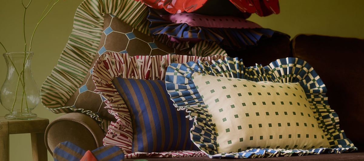 Piecework pillows with ruffled, scalloped, and checkered trims piled on a brown velvet sofa against an olive wall