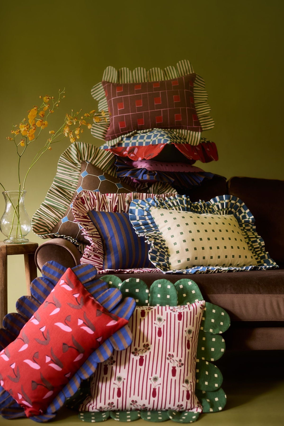 A stack of Piecework pillows with ruffled and scalloped trims on a brown velvet sofa beside a vase of yellow flowers