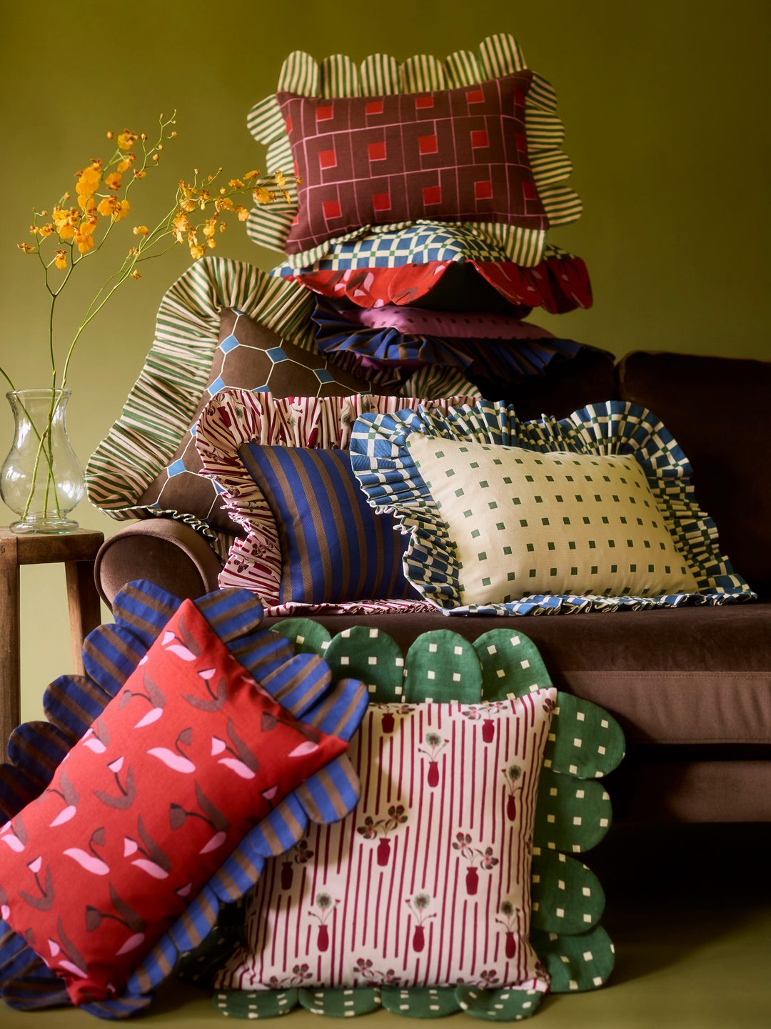 Piecework petal and ruffle pillows piled high on a brown velvet sofa beside a vase of yellow orchids