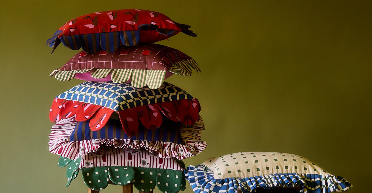A tall stack of Piecework petal, ruffle and scalloped pillows in red, blue, pink and green prints on a wooden stool against an olive green wall